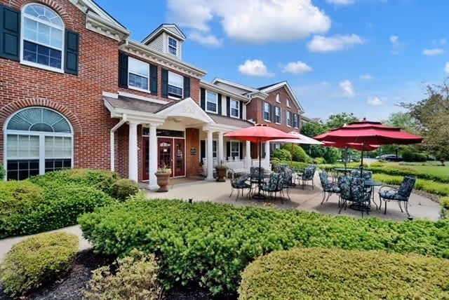 Exterior view of a two-story brick building with white trim and large windows. The entrance has a covered porch with columns and the number 500 above the door. In front of the building, there is a patio area with several tables and chairs under red umbrellas. Well-maintained green bushes and landscaping surround the patio and building. The sky is blue with a few clouds.
