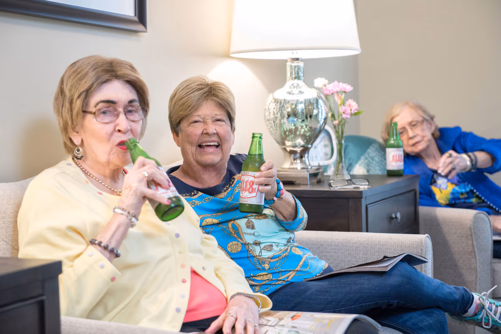 Three elderly women sitting comfortably in a living room area, each holding a green bottle of Ale-8-One soda. They are smiling and appear to be enjoying a relaxed moment together. The room has a lamp, a side table with flowers, and magazines.