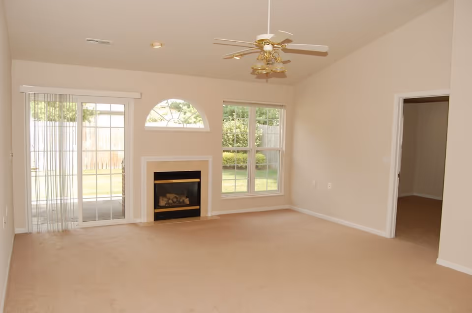 Empty living room with beige carpet, a ceiling fan with lights, a fireplace, a large window, a sliding glass door with vertical blinds, and an open doorway leading to another room.