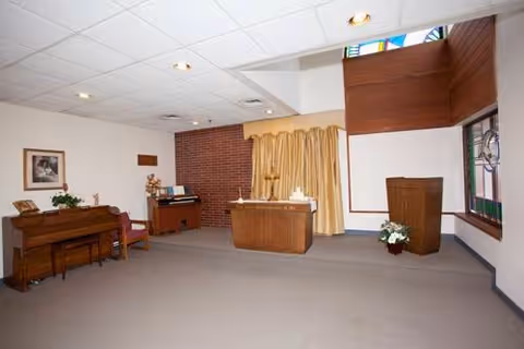 Interior room with a piano, organ, wooden podium, and altar-like table with candles, featuring a brick wall, beige curtains, and stained glass windows.