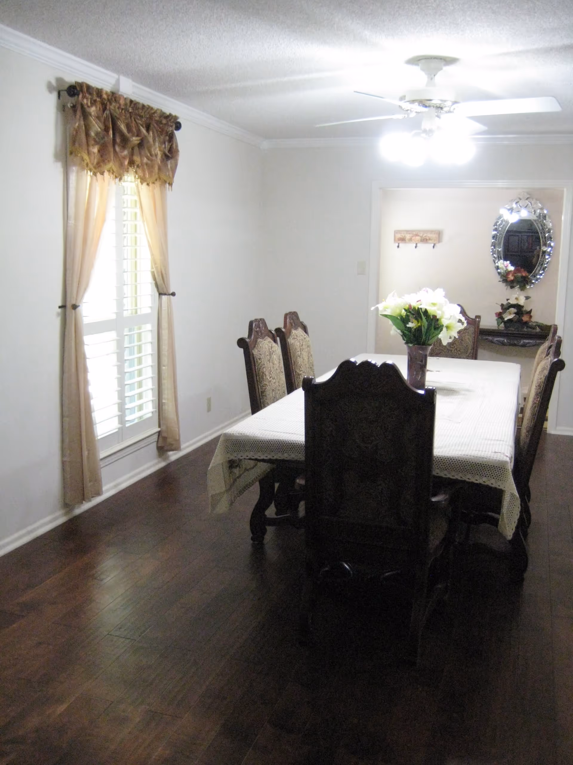A dining room with a long wooden table covered with a white lace tablecloth and six ornate wooden chairs. A vase with white flowers is placed in the center of the table. The room has dark hardwood floors, a window with beige curtains on the left wall, and a ceiling fan with lights above the table. In the background, there is a decorative mirror and a small table with flowers on it.