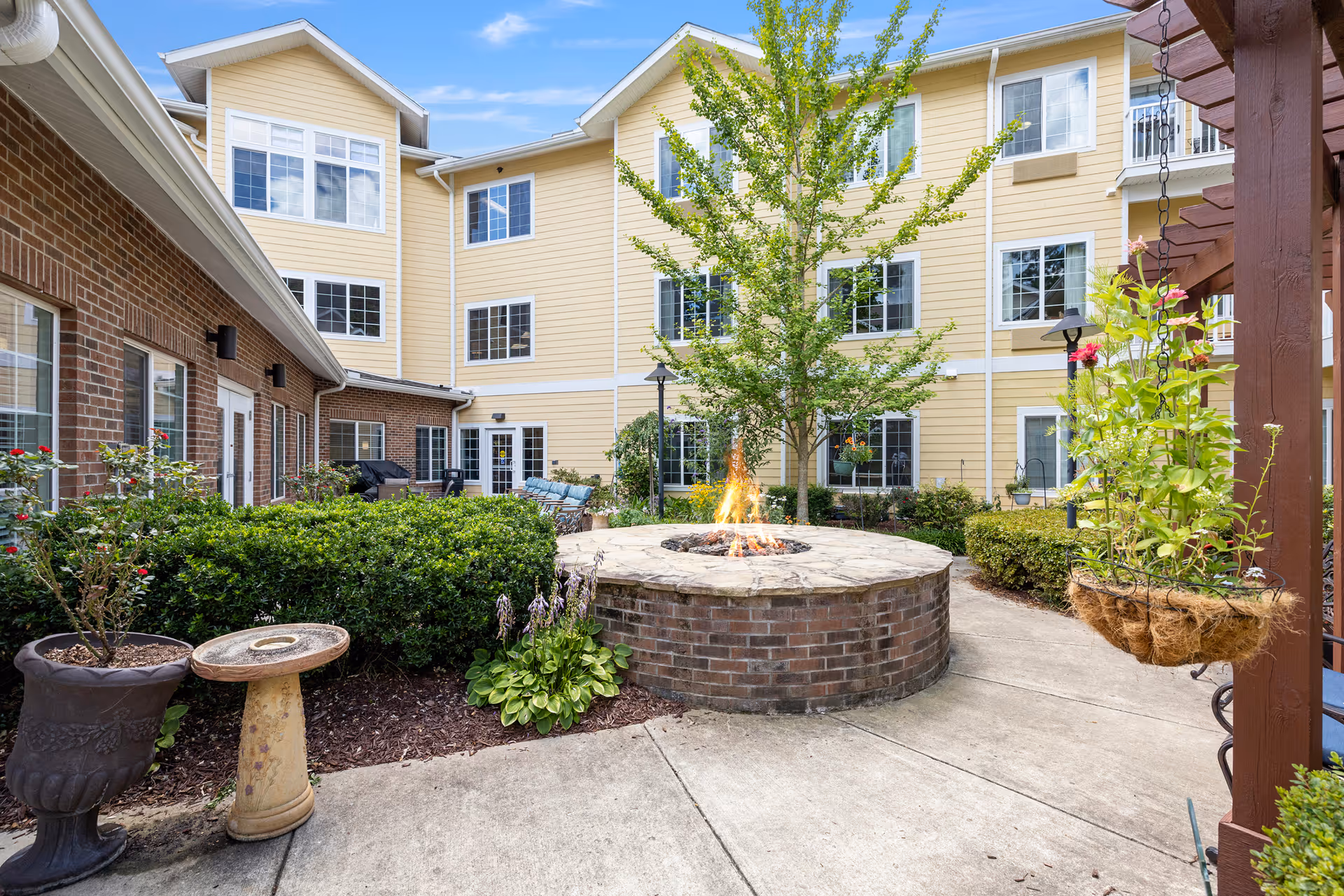Outdoor courtyard area of a senior living facility with a circular brick fire pit in the center, surrounded by greenery, potted plants, and a tree. The courtyard is enclosed by a three-story building with yellow siding and white-trimmed windows. There is a hanging plant on the right side under a wooden pergola.