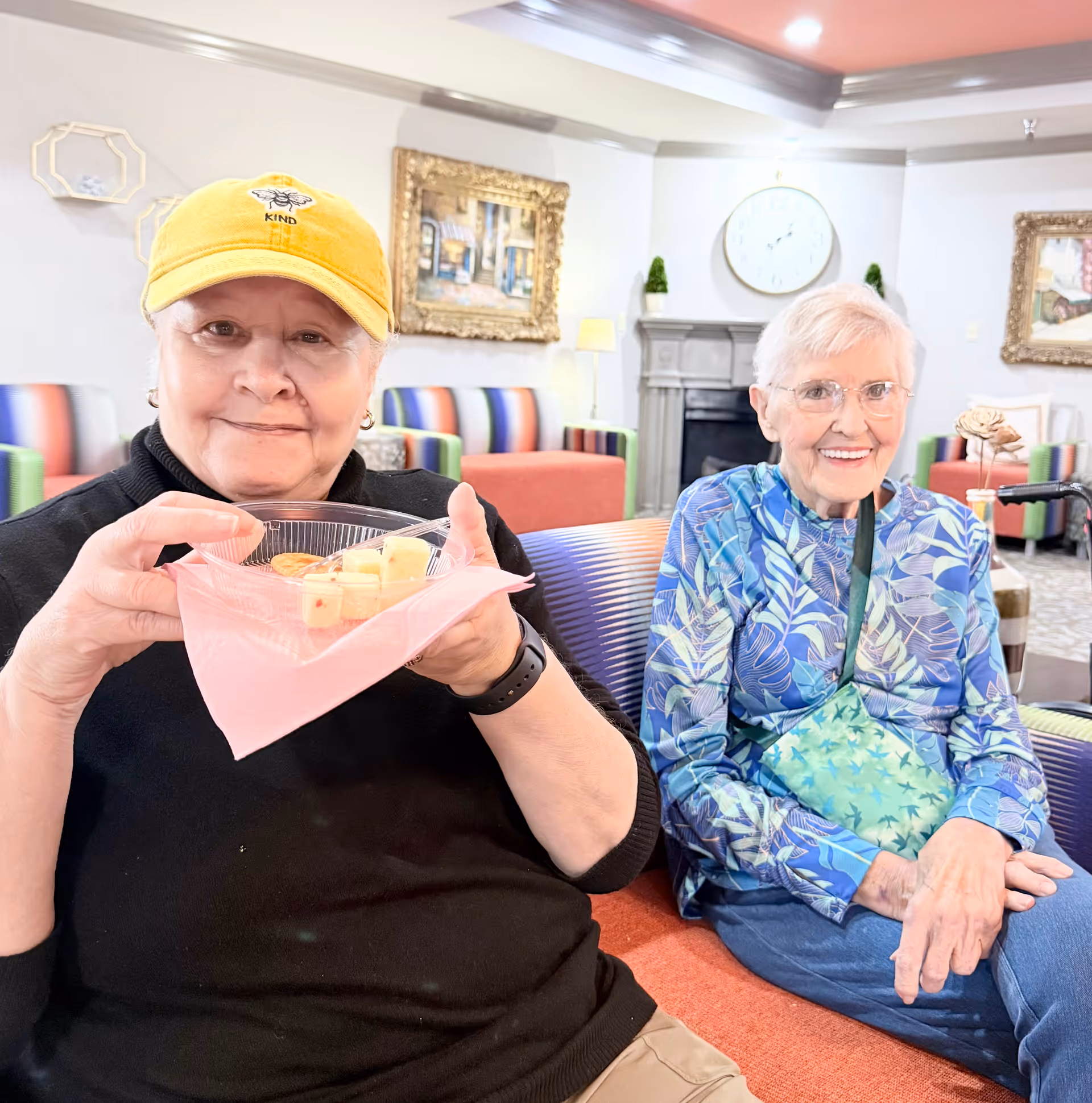 Two elderly women sitting on a colorful striped couch in a well-lit living room. One woman wearing a yellow cap and black sweater is holding a clear plastic bowl with fruit and a pink napkin. The other woman, wearing glasses and a blue patterned shirt, is smiling. The background shows a fireplace, a large clock, framed paintings, and more colorful furniture.