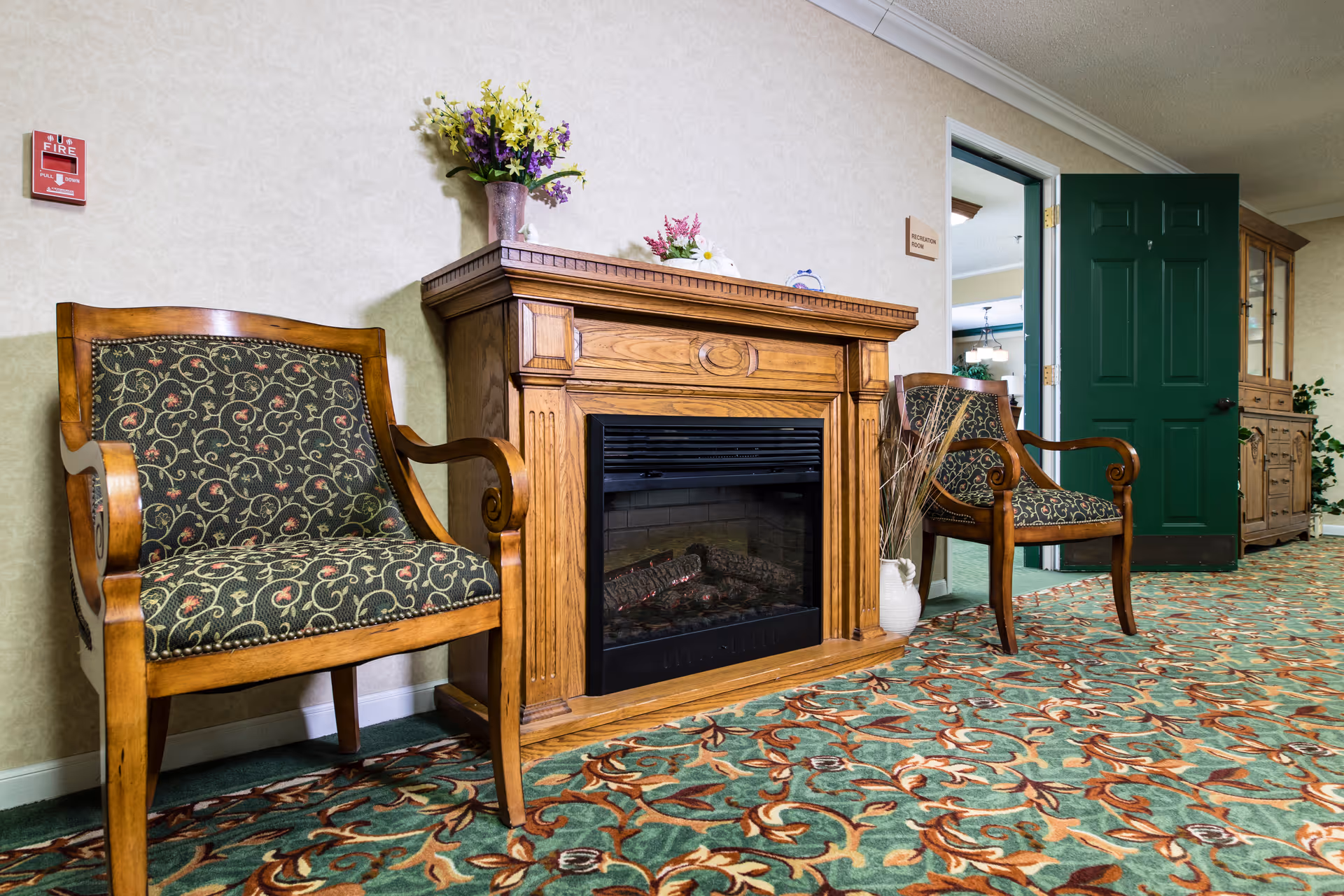 Two ornate wooden armchairs flanking a decorative electric fireplace in a carpeted common area.