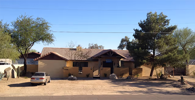 Single-story building with a tiled roof and beige exterior walls, surrounded by trees and desert landscaping, with a car parked in front on a dirt driveway under a clear blue sky.