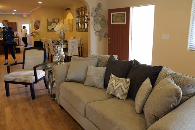 Interior view of a senior living facility showing a beige sofa with multiple cushions and a matching armchair in the foreground. In the background, there is a dining area with a table and chairs, decorative wall art, and a few people standing and conversing. The room has wooden flooring and light-colored walls with a red front door visible.