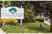 Outdoor view of the entrance area of Brinton Woods Nursing & Rehabilitation Center at Winfield, featuring a white sign with the facility name surrounded by green bushes and trees, with a bench and a building visible in the background.