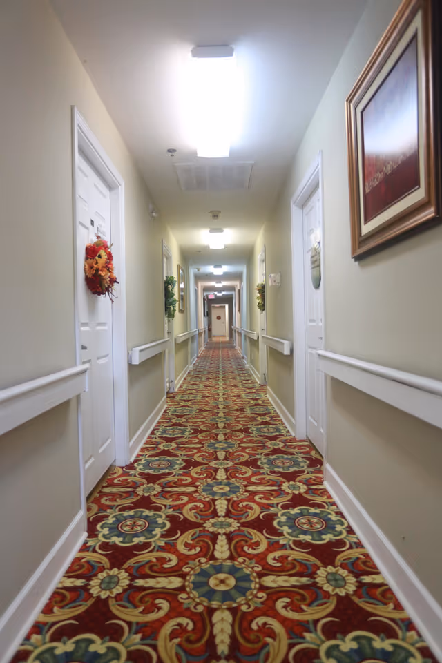 A long, narrow hallway in a senior living facility with patterned red and gold carpet, beige walls, white doors decorated with wreaths, handrails on both sides, ceiling lights, and framed artwork on the walls.