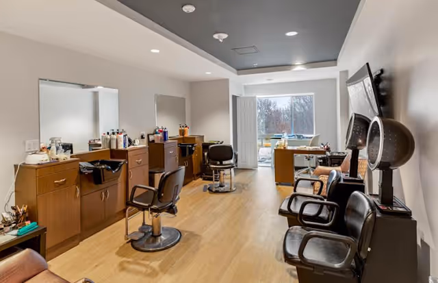 Interior view of a salon area in a senior living facility with wooden flooring, salon chairs, mirrors, and hair care products on wooden cabinets. There is a seating area with black chairs and a TV mounted on the wall.
