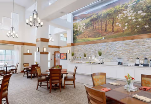 A spacious and well-lit dining room with several wooden tables and chairs arranged neatly. The room features high ceilings with modern chandeliers, large windows allowing natural light, and a long buffet counter against one wall. Above the buffet is a large mural depicting a vineyard landscape. The decor is warm and inviting, with neutral tones and subtle accents.
