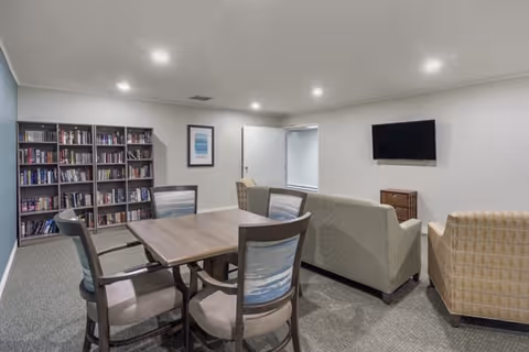 A cozy common area in a senior living facility featuring a wooden table with four cushioned chairs, two sofas, and an armchair arranged around a wall-mounted flat-screen TV. A bookshelf filled with books and framed artwork are visible against the walls.