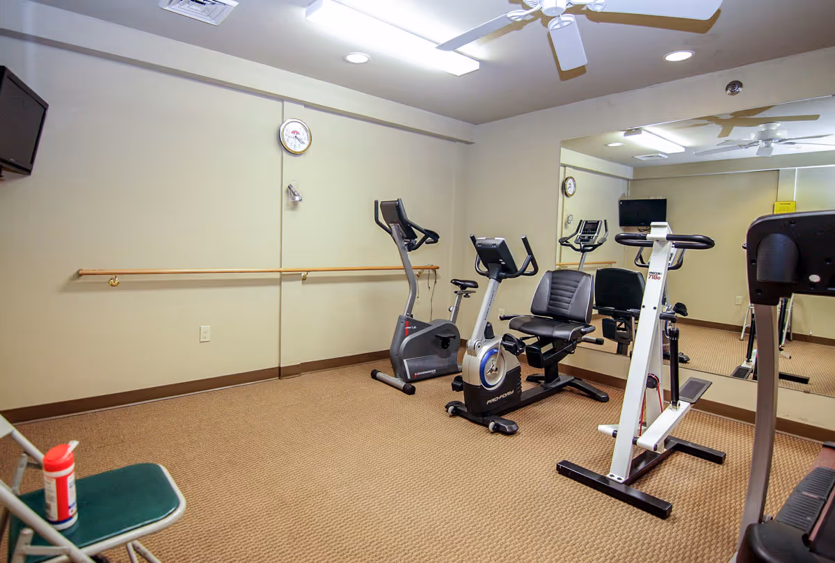 Small exercise room with beige walls and carpet, containing stationary exercise bikes and other fitness equipment. A large mirror covers one wall, reflecting the equipment and a wall clock. A folding chair with a container of disinfectant wipes is placed in the corner.