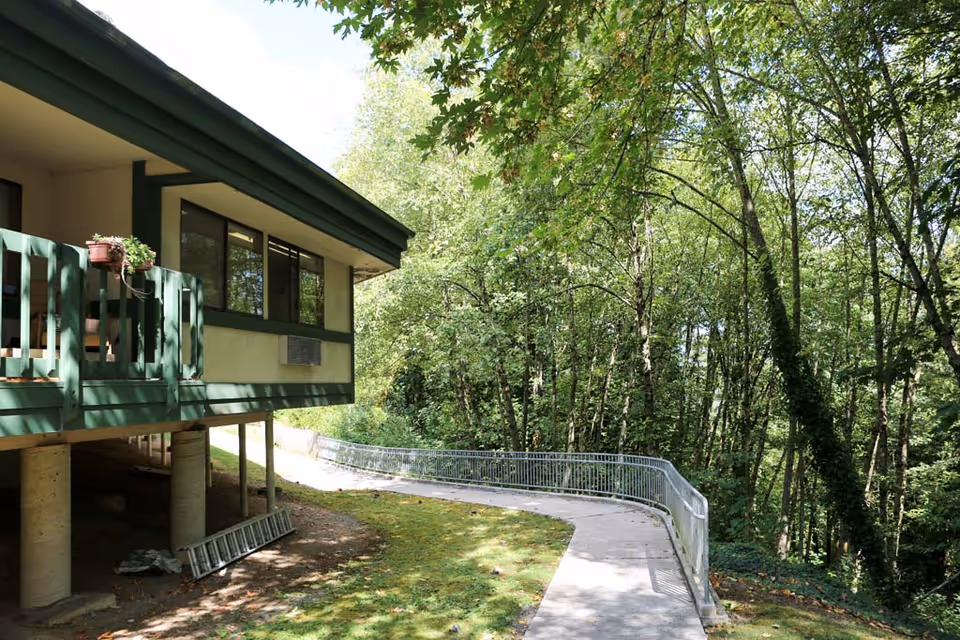 Exterior view of a building with green trim and a small balcony with potted plants. A concrete pathway with a metal railing curves alongside the building, surrounded by lush green trees and grass.