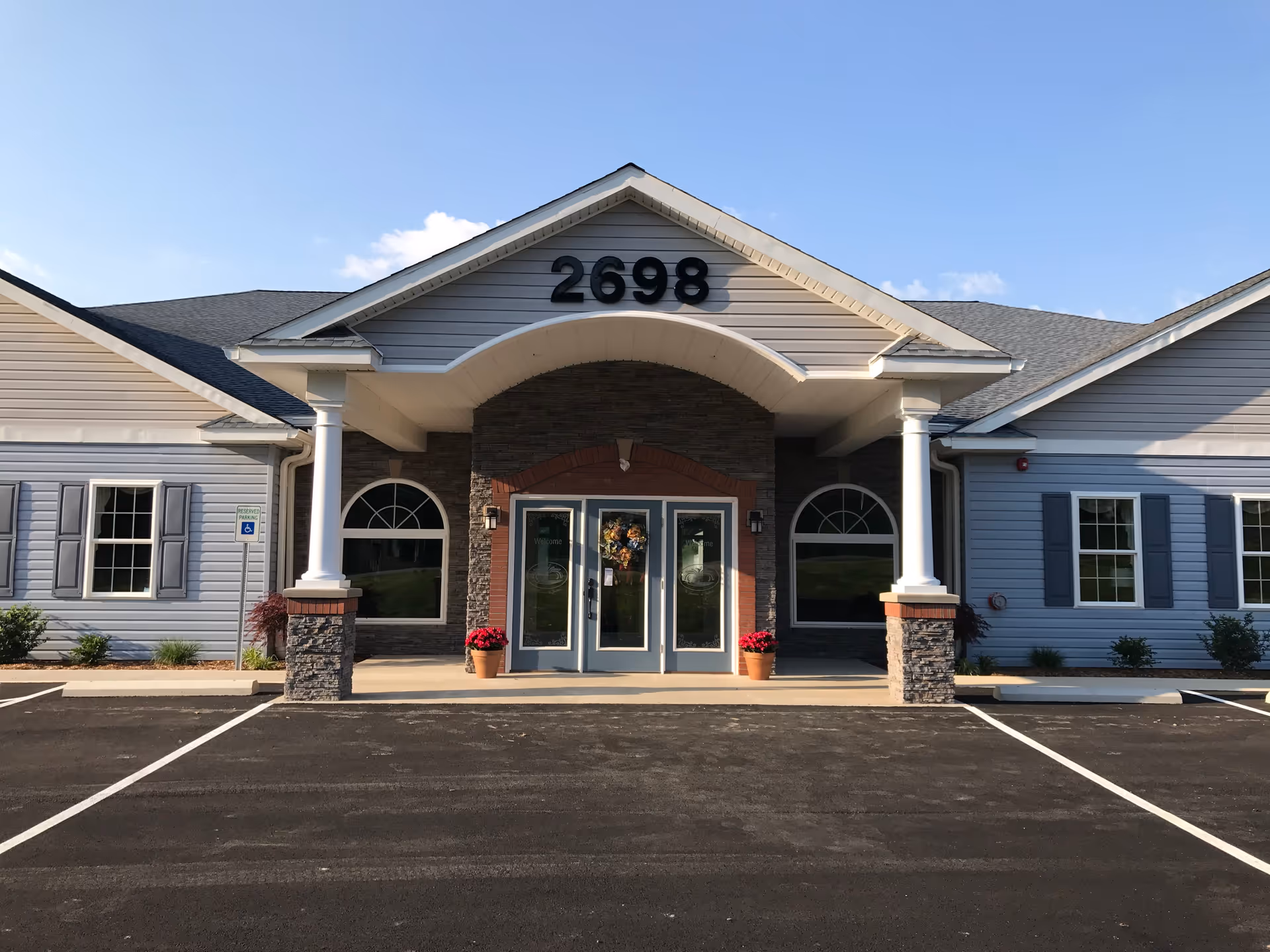 Entrance of a single-story light-gray building with address number 2698 above a covered portico, double glass doors, two columns, and a parking area in front.