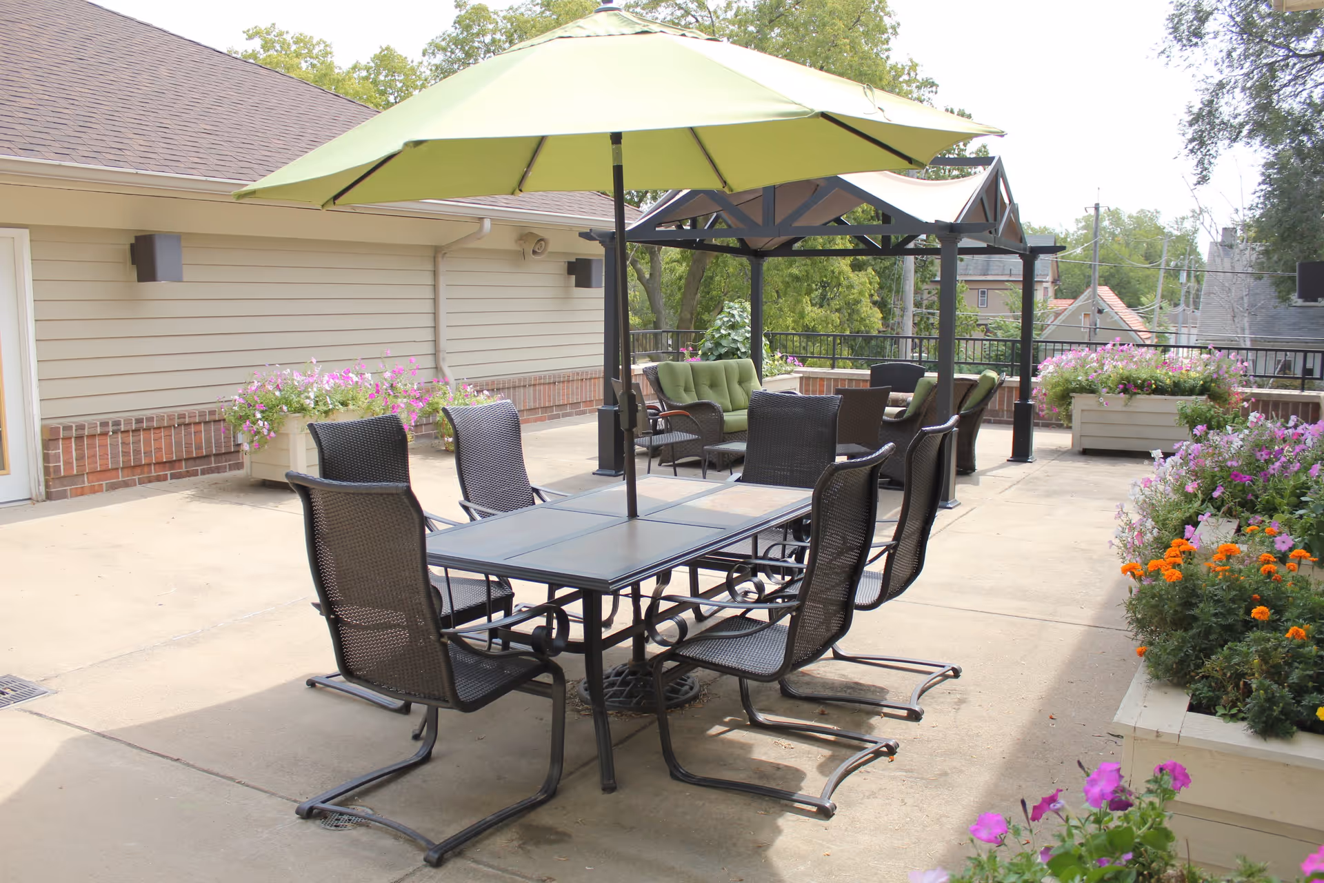 Outdoor patio area with a rectangular table and six black metal chairs, shaded by a large green umbrella. In the background, there is a seating area with green cushioned chairs under a black pergola. The patio is surrounded by planters filled with colorful flowers and greenery.