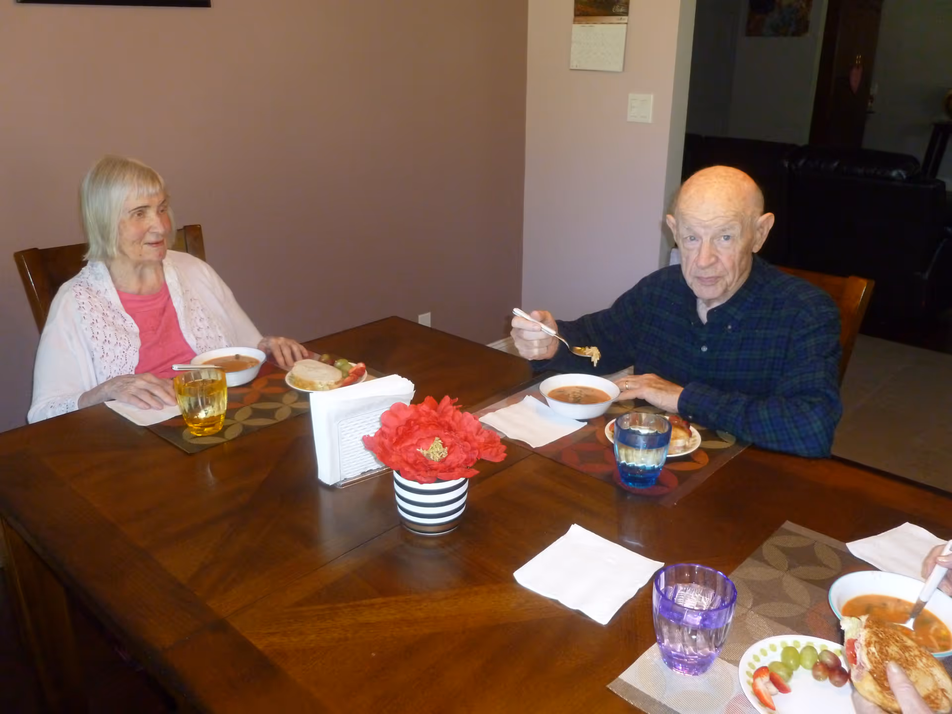 Two elderly individuals sitting at a wooden dining table eating soup. The woman on the left is wearing a pink shirt and white cardigan, and the man on the right is wearing a dark plaid shirt. The table has placemats, glasses of water, a napkin holder, and a red flower centerpiece.