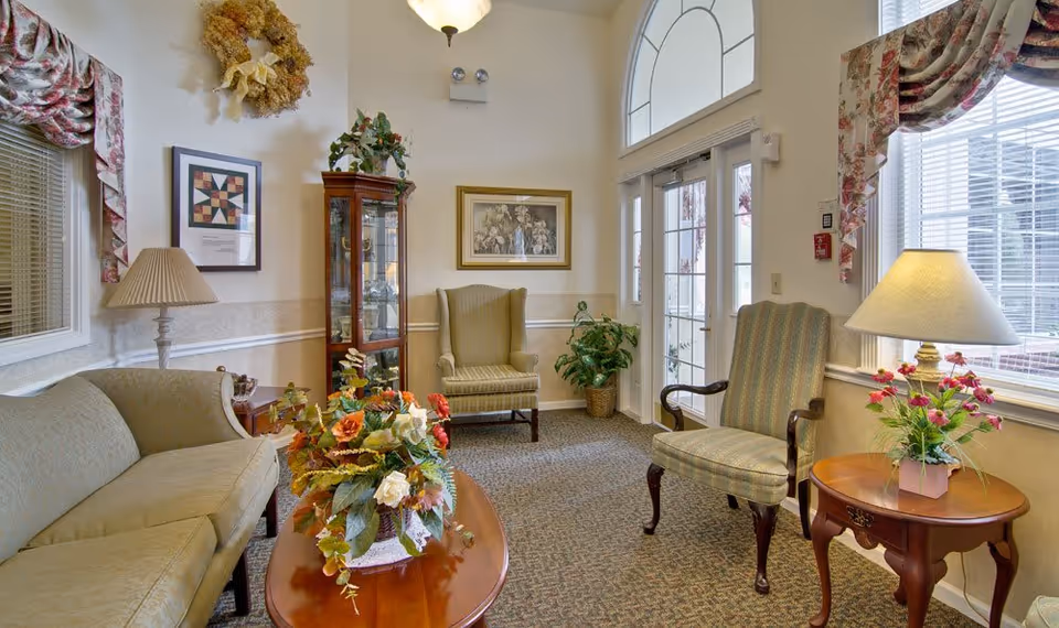 A cozy living room area with a beige sofa, two armchairs, a wooden coffee table with a floral arrangement, a side table with a lamp and flowers, a glass display cabinet, framed artwork on the walls, and large windows with floral curtains letting in natural light.