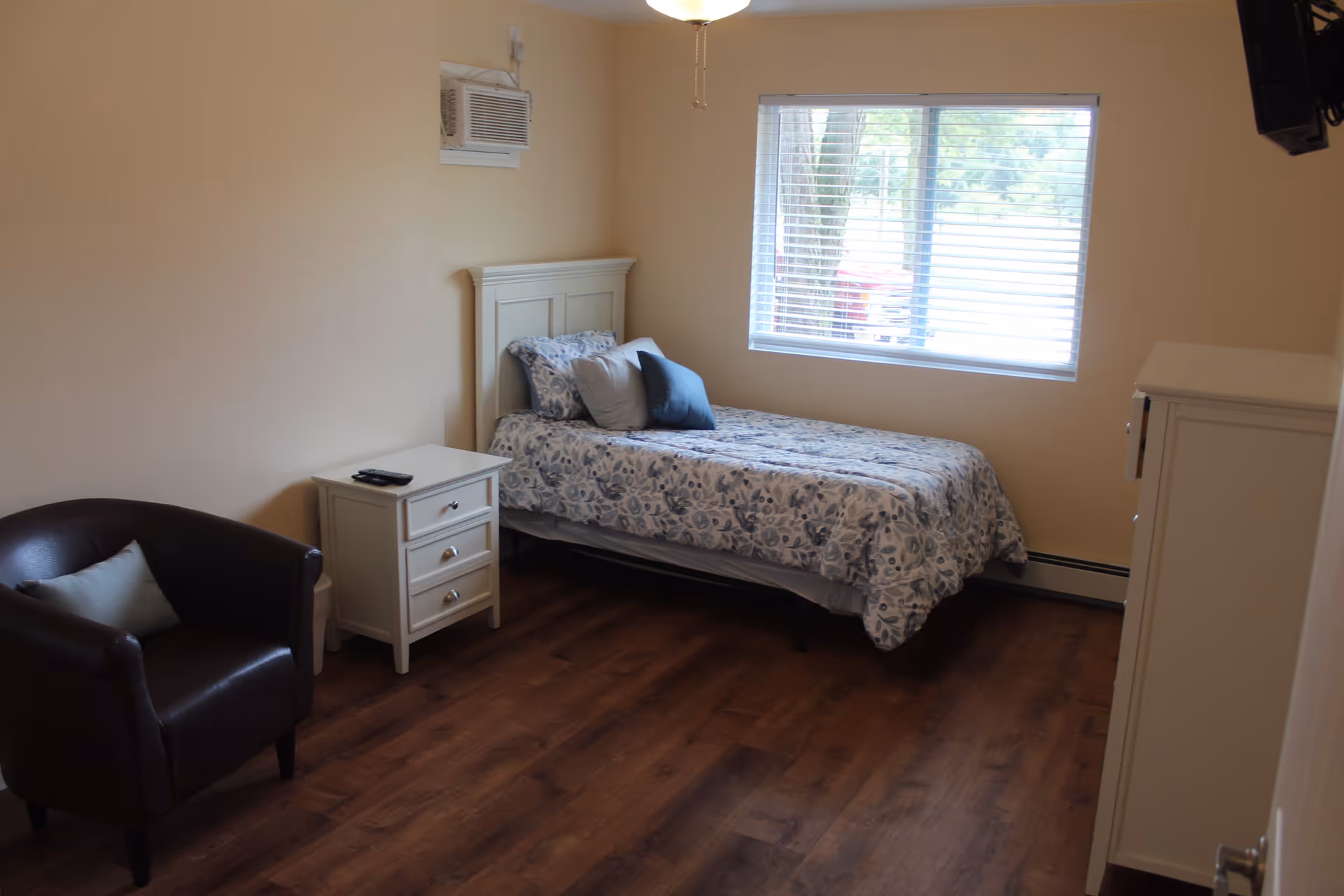 A simple bedroom with a single bed covered in a floral patterned comforter and three pillows. Next to the bed is a white nightstand with a remote control on top. There is a dark brown armchair with a light blue pillow on it. The room has wood flooring, beige walls, a window with blinds letting in natural light, and a wall-mounted air conditioning unit above the bed.