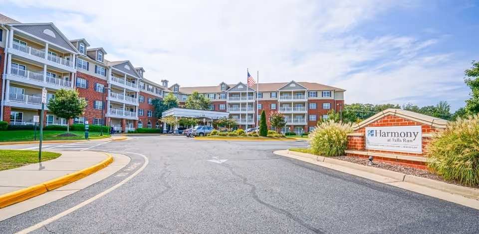Exterior view of Harmony at Falls Run, a multi-story senior living facility with red brick and beige siding, balconies, and an American flag flying in front. The driveway leads to the entrance with a covered drop-off area, and a landscaped sign with the facility's name is visible on the right.