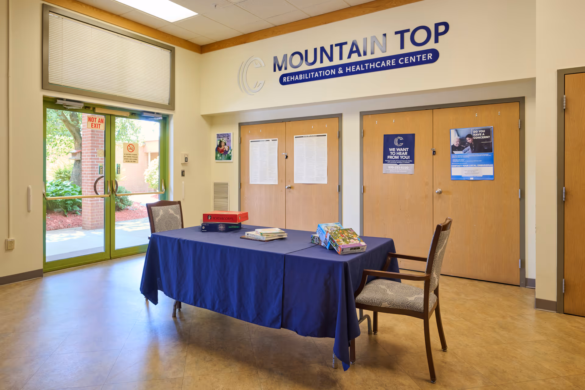 Interior view of the Mountain Top Rehabilitation and Healthcare Center lobby or common area with a table covered in a blue tablecloth, two chairs, informational posters on wooden doors, and a glass exit door showing greenery outside.