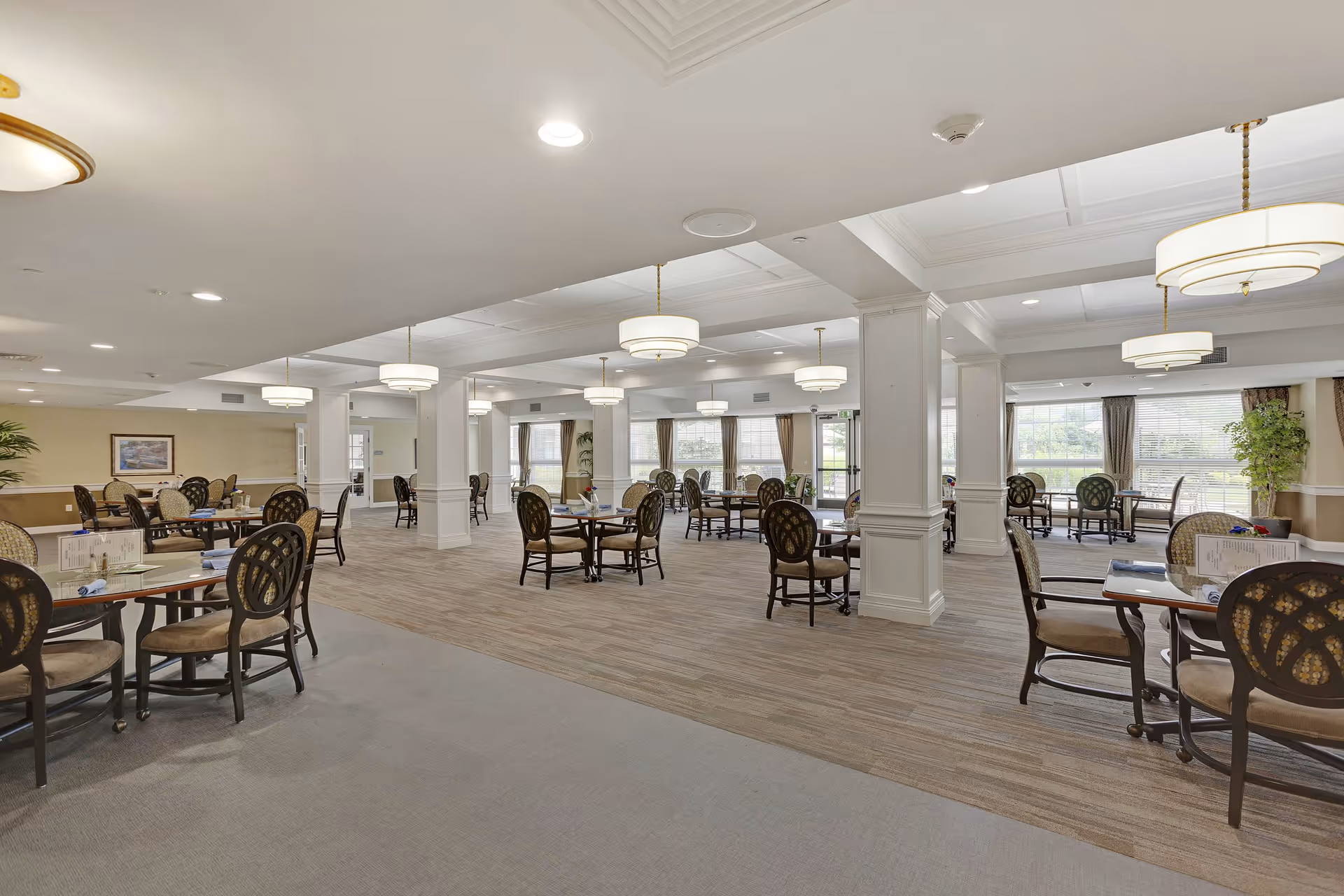 A spacious dining room in a senior living facility with multiple round tables and chairs arranged neatly. The room features large windows with curtains allowing natural light to fill the space, white columns, and modern ceiling lights. The floor is carpeted with a combination of solid and patterned sections.