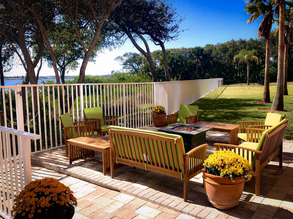 Outdoor patio area with wooden seating furniture featuring green cushions arranged around a central fire pit table. The patio is paved with bricks and decorated with potted yellow flowers. A white metal fence borders the patio, with trees and a body of water visible in the background under a clear blue sky.