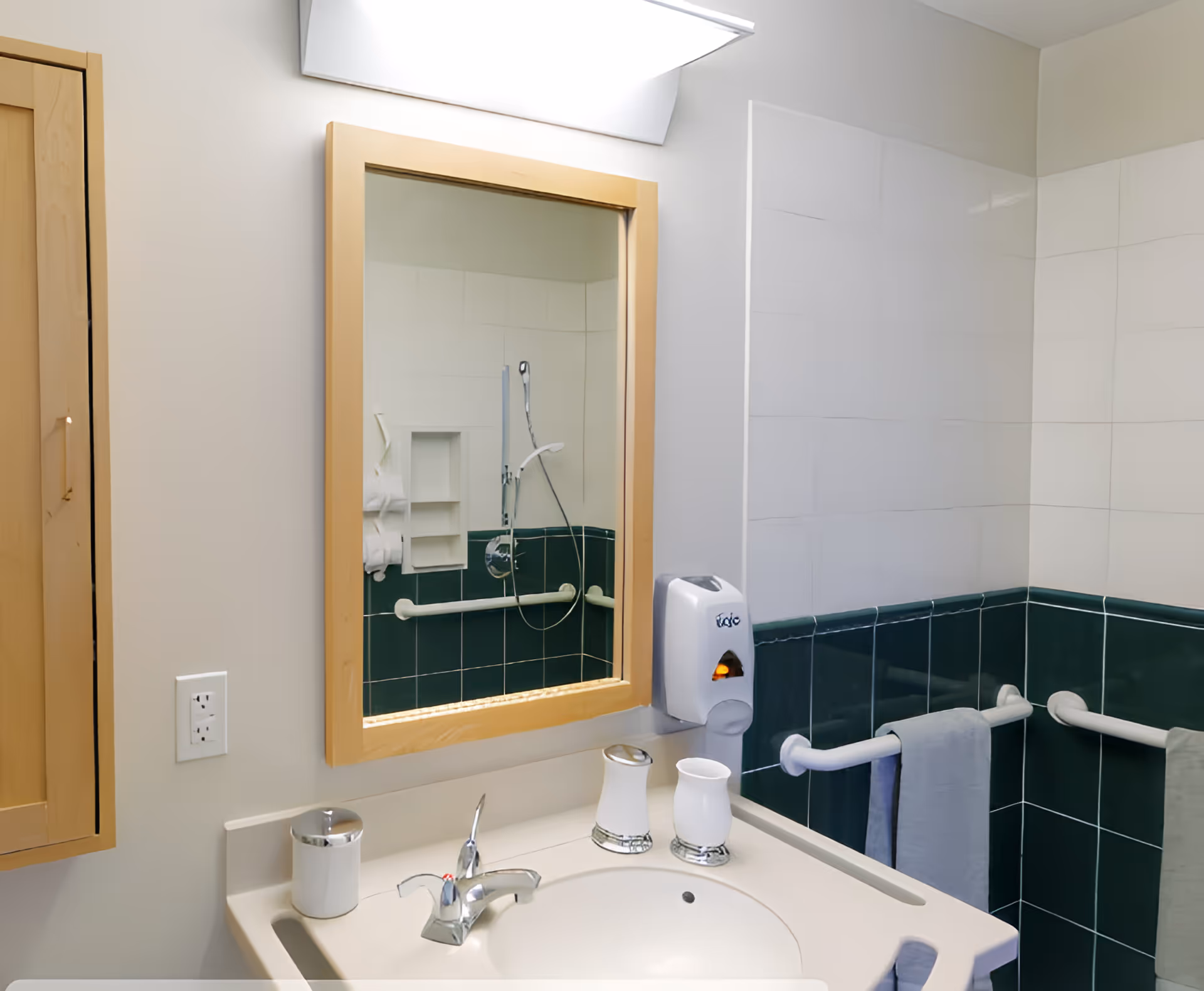 A bathroom sink area with a beige countertop, a silver faucet, a soap dispenser, and two white containers. Above the sink is a wooden-framed mirror reflecting a shower area with white and dark green tiles, a handheld showerhead, and grab bars. There are also grab bars with towels hanging on them on the right side of the image.