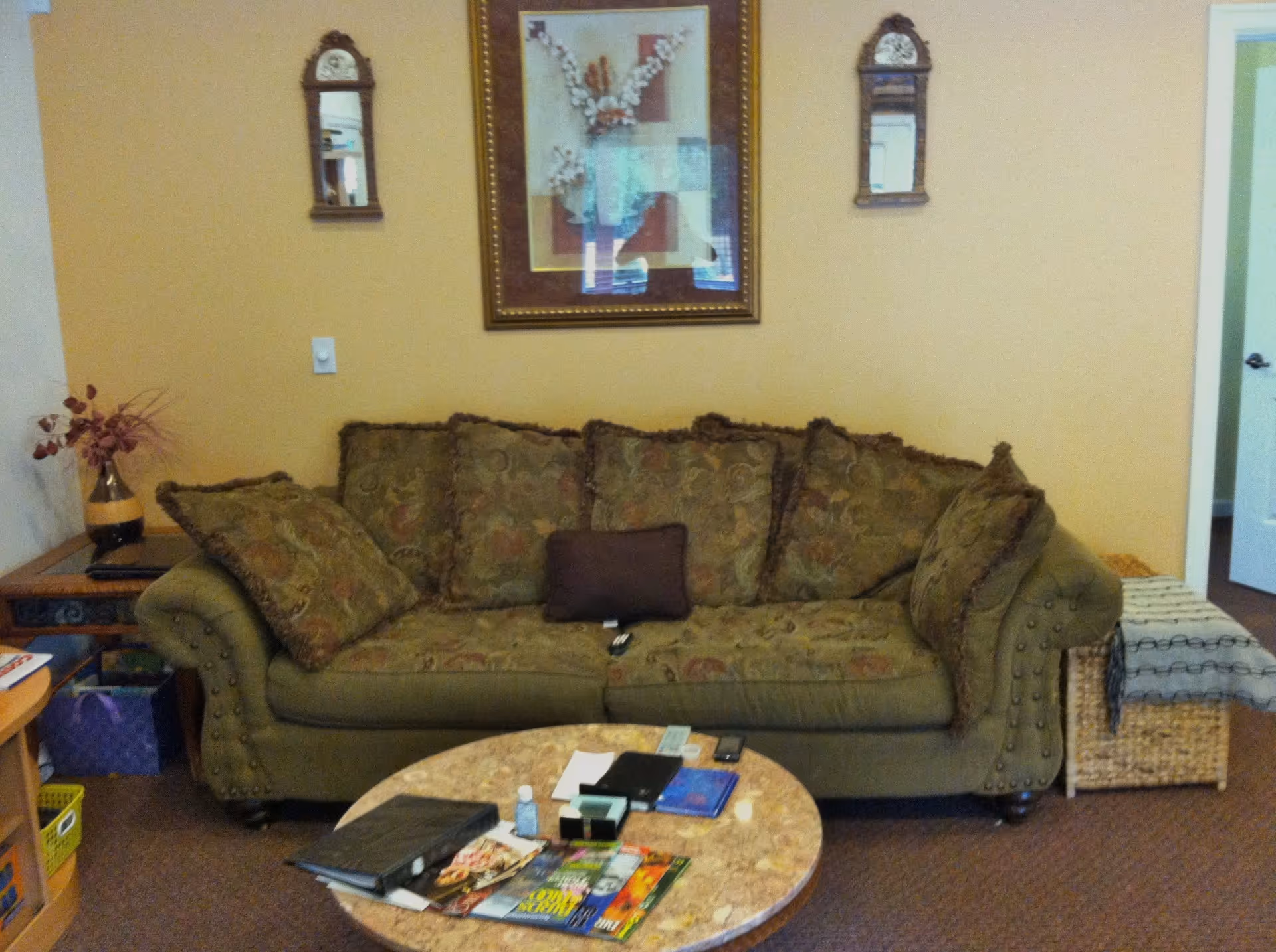 A cozy living room area with a green patterned sofa adorned with multiple cushions. Above the sofa hangs a framed floral artwork flanked by two small decorative mirrors. In front of the sofa is a round marble-top coffee table cluttered with magazines, a binder, a small bottle, and other miscellaneous items. To the left of the sofa is a side table with a vase of dried flowers, and to the right is a woven basket with a folded blanket on top. The walls are painted a warm beige color.