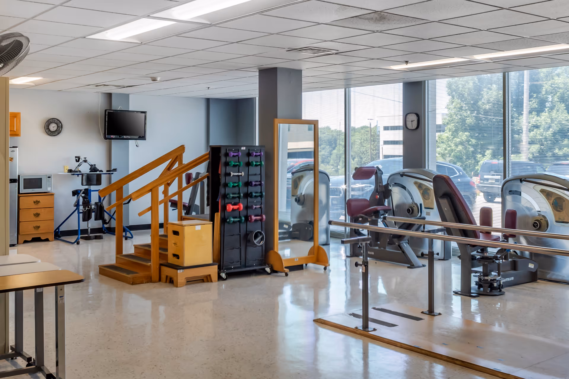 A rehabilitation and exercise room with large windows showing parked cars outside. The room contains exercise equipment including stationary bikes, a set of colorful dumbbells on a rack, a wooden staircase for physical therapy, parallel bars for walking practice, a full-length mirror, a wall-mounted TV, and a small wooden cabinet with a microwave on top.