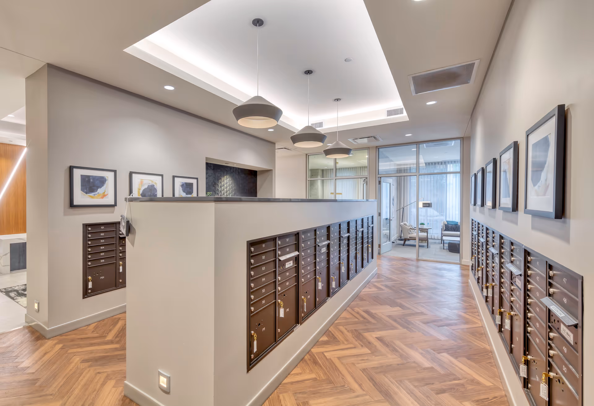 Interior view of a modern mailroom area in a senior living facility with rows of dark brown mailboxes on both sides of a central partition. The room features wood-patterned flooring, recessed ceiling lights, three pendant lights hanging from a tray ceiling, and framed abstract artwork on the walls. A glass door leads to a seating area with chairs and a table in the background.