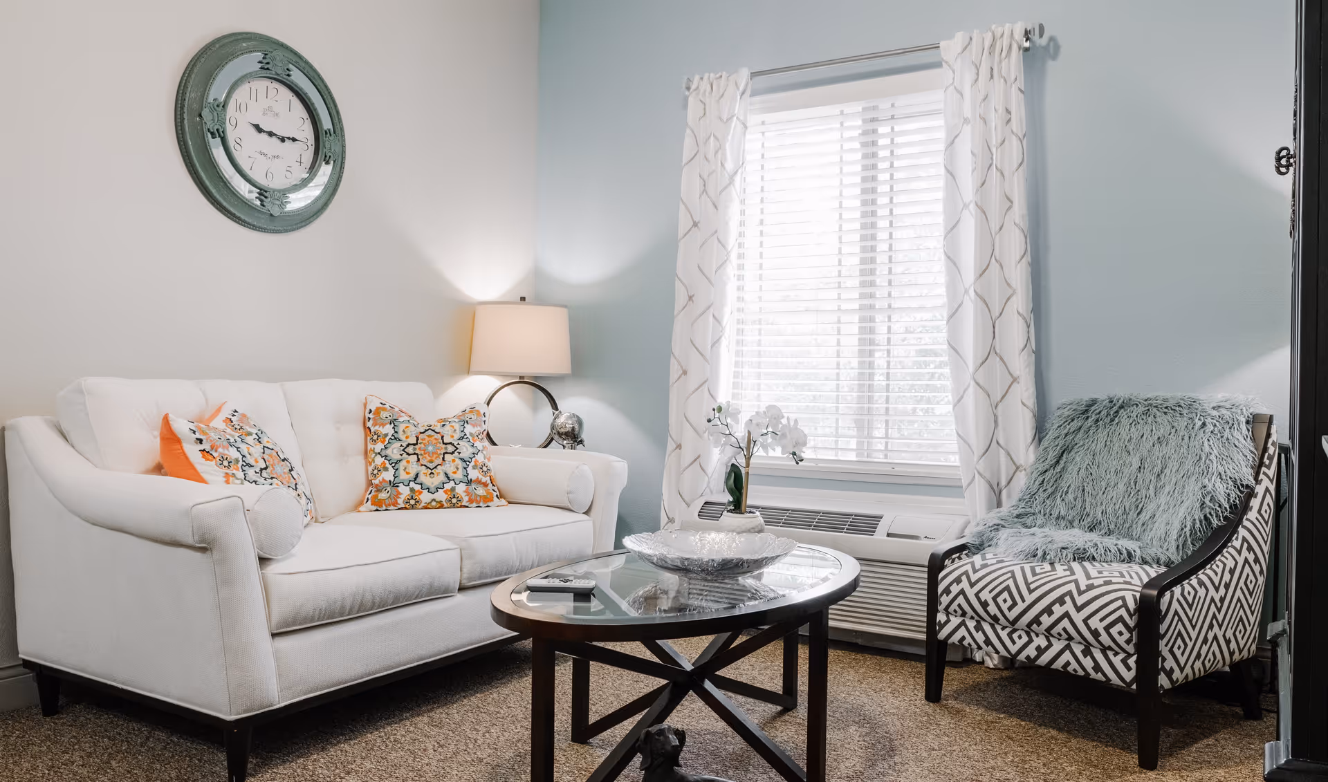 A cozy living room with a white couch adorned with colorful patterned pillows, a patterned armchair with a fluffy blue throw, a round glass coffee table with a decorative bowl and a small vase with flowers, a window with white blinds and patterned curtains, a wall clock, and a table lamp on a side table.