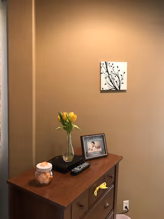 A wooden dresser against a beige wall with a small framed photo, a glass vase with yellow tulips, a remote control, and a jar with orange and white items on top. A small black and white artwork featuring a branch with leaves is hung on the wall above the dresser.