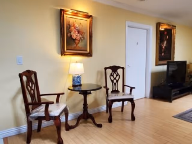 Two wooden armchairs flank a small round table with a lamp against a yellow wall, with framed paintings and a TV on a low stand in the background.