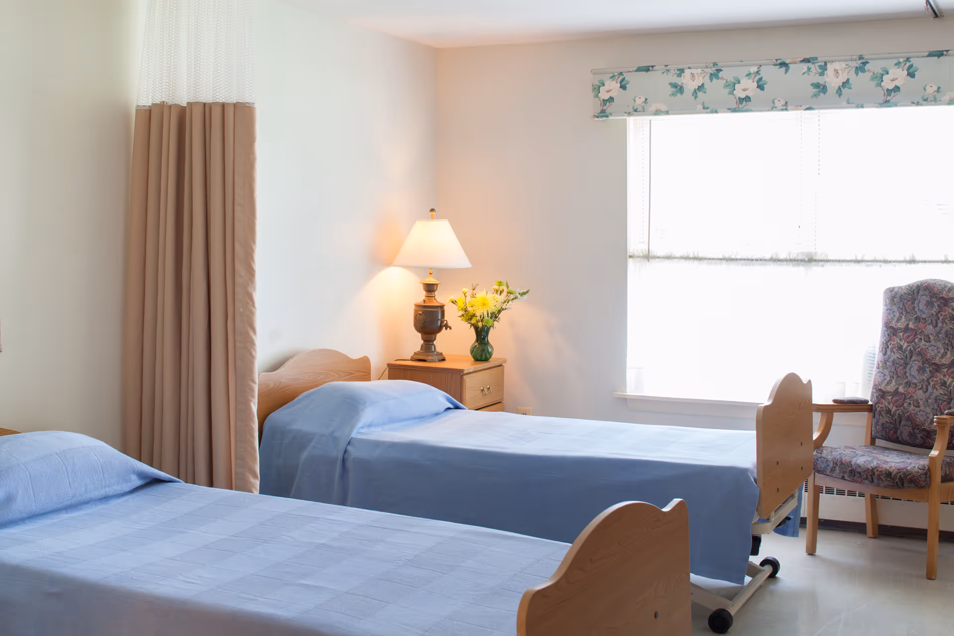 A bright room with two single beds covered in light blue bedspreads, a wooden nightstand with a lamp and a vase of yellow flowers, a floral-patterned armchair near a window with white blinds and a floral valance, and a beige privacy curtain partially visible on the left.
