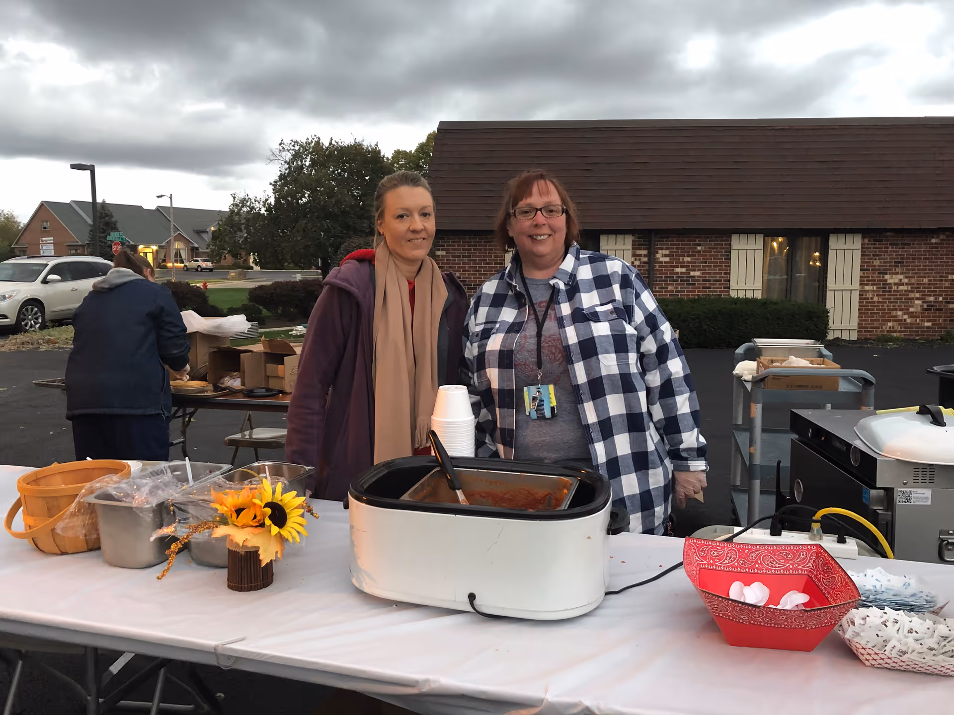 Two women standing behind a table set up outdoors with food containers, a slow cooker, disposable cups, and a decorative sunflower. One woman is wearing a purple jacket and beige scarf, and the other is wearing a checkered shirt and glasses. In the background, there is a brick building and another person working at a table with boxes.