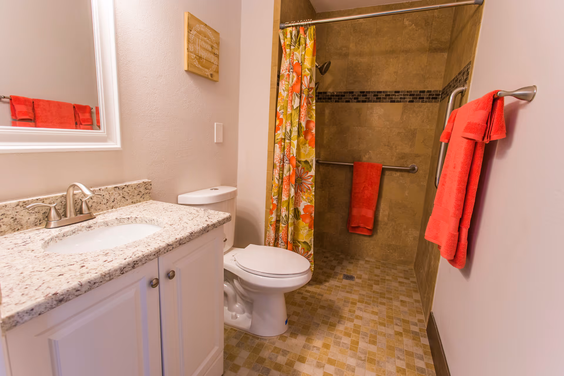 A clean and well-lit bathroom featuring a white vanity with a granite countertop and a sink, a toilet, and a walk-in shower with beige tiles and a floral shower curtain. Red towels hang on a towel rack and a grab bar is installed inside the shower for safety.