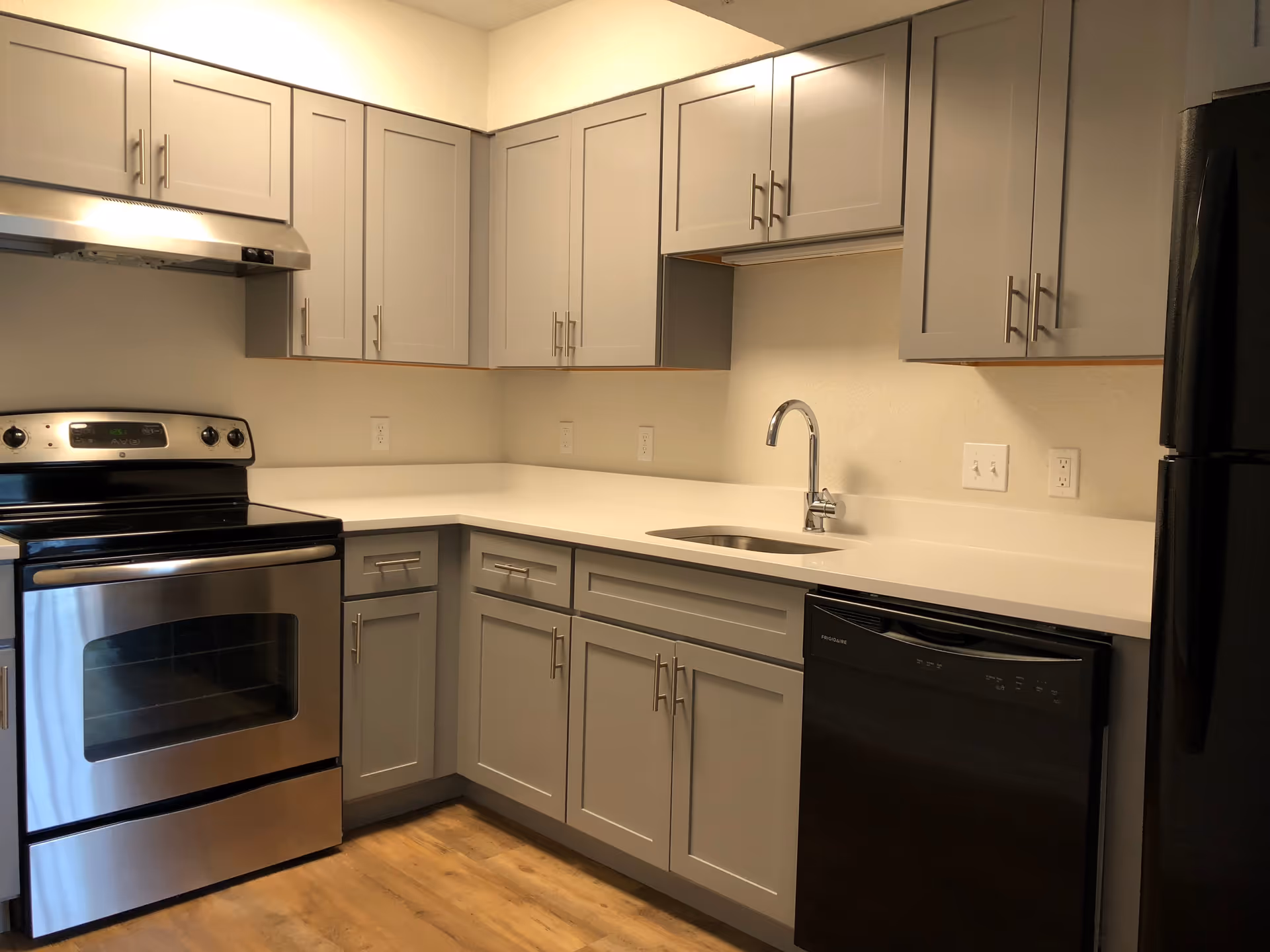 Modern kitchen with gray cabinets, a stainless steel oven and range hood, a black dishwasher, a black refrigerator, a white countertop, and a stainless steel sink with a curved faucet. The floor is wood-style and the walls are light-colored.