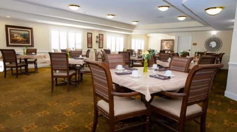 A dining room with multiple tables covered in beige tablecloths, each set with white cups, saucers, and napkins. The chairs have wooden frames with striped cushions. The room is well-lit with ceiling lights and natural light from windows. There are framed artworks on the walls and floral centerpieces on the tables.