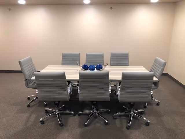 A modern conference room with a rectangular light wood table surrounded by eight gray office chairs on wheels. The table has a decorative centerpiece with blue spherical objects. The room has beige walls and carpeted floor with ceiling lights.
