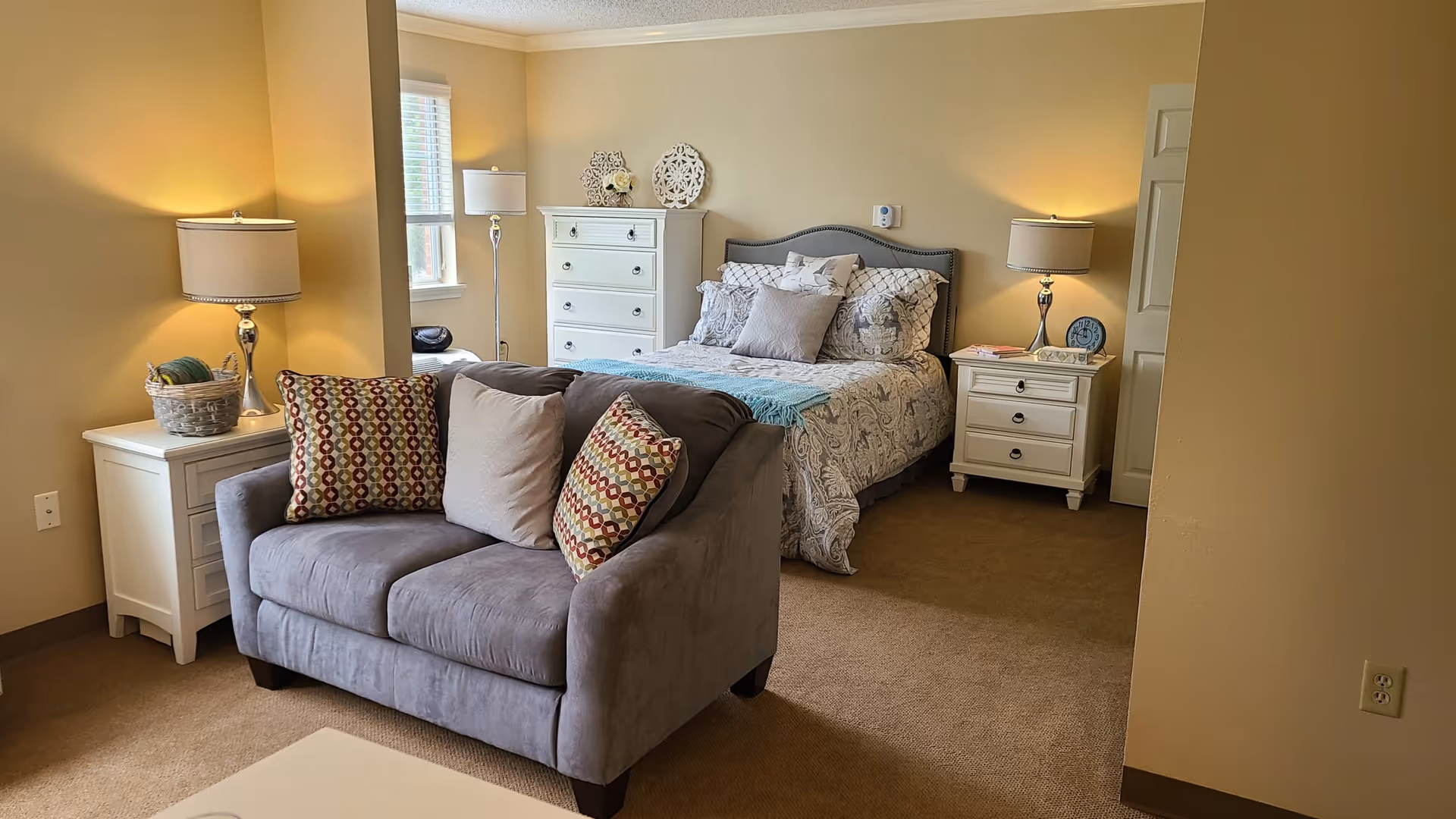 A cozy bedroom with a neatly made bed featuring patterned bedding and multiple pillows. There are two white nightstands on either side of the bed, each with a lamp. A white chest of drawers is positioned against the wall near a window with blinds. In front of the bed is a small gray loveseat with three decorative pillows. The room has beige walls and carpeted flooring.