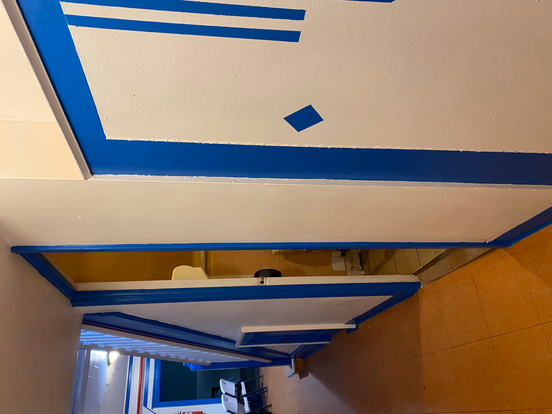 Interior hallway of a facility with blue and white painted walls and trim. There is an open door revealing a small bathroom with a sink and tiled walls. The hallway has a staircase with white railings and some white folding chairs are visible in the background.