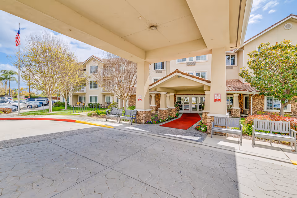 Covered front entrance of a senior living building with a red carpet, benches, and landscaped grounds.