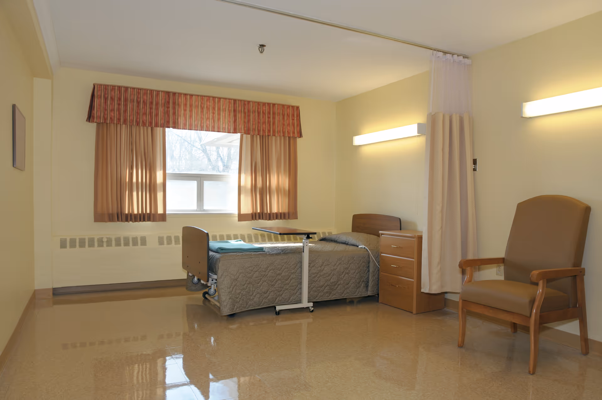 A simple, clean room with a single hospital-style bed covered with a gray quilt, a wooden bedside table with drawers, a brown armchair, and a window with beige curtains and a red valance. The room has beige walls and a shiny floor, with two horizontal wall lights and a privacy curtain partially drawn.