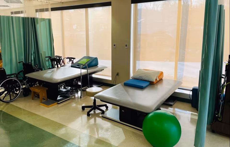 Physical therapy room with two treatment tables, a green exercise ball, wheelchairs, stools, and green privacy curtains. Large windows with beige blinds allow natural light into the room.