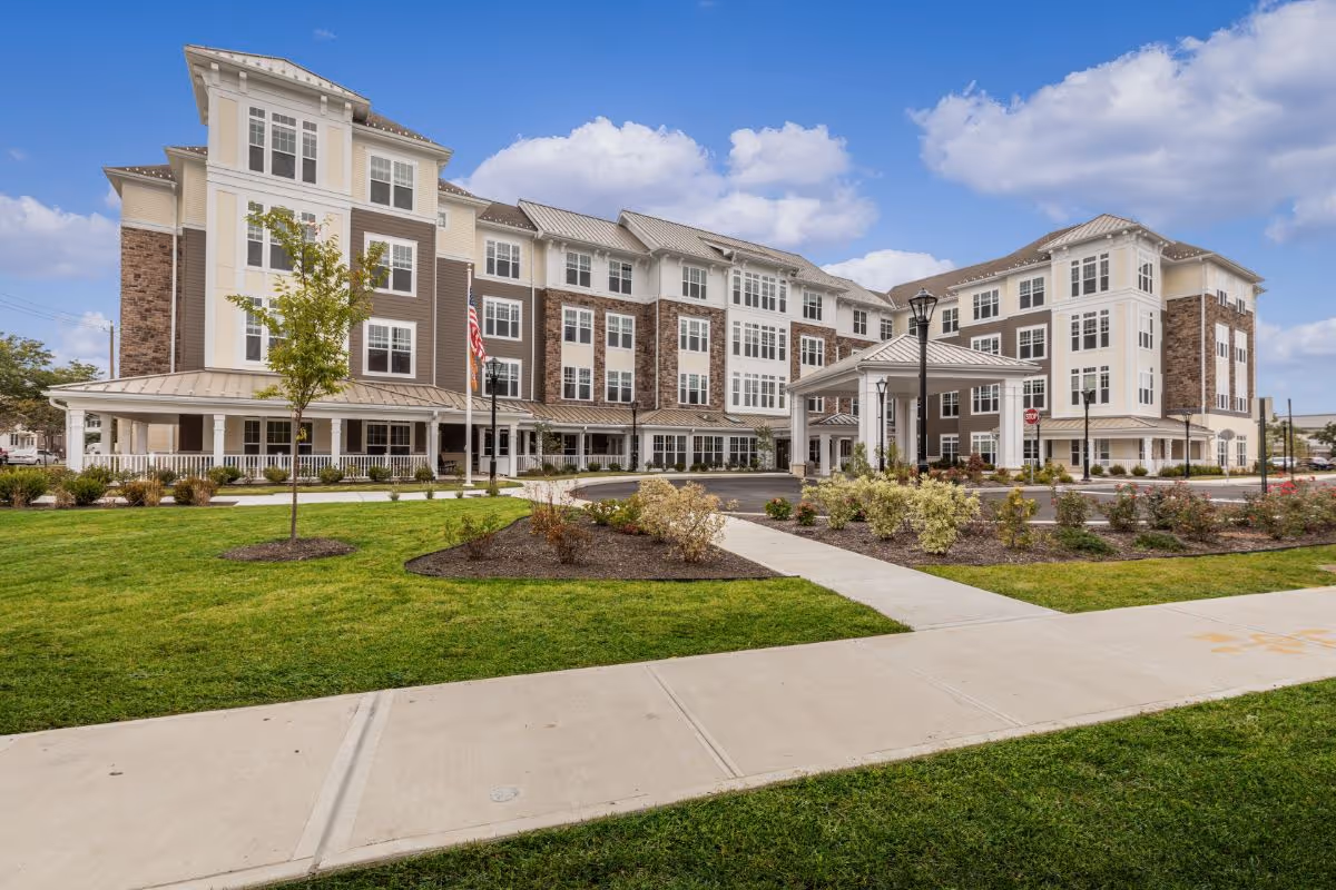 Exterior view of a large, modern senior living facility building with multiple floors, large windows, and a covered entrance. The building is surrounded by well-maintained green lawns, landscaped bushes, and a paved walkway under a partly cloudy sky.