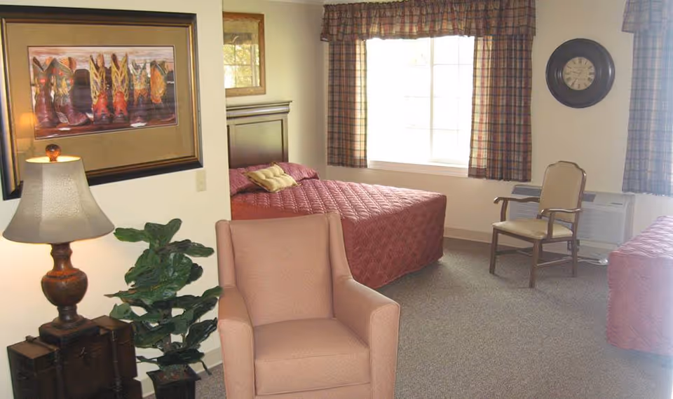 A bedroom in a senior living facility with two beds covered in maroon quilted bedspreads, a cushioned armchair in the foreground, a wooden chair near a window with plaid curtains, a wall clock, a table lamp on a small chest, a green potted plant, and a framed picture of colorful cowboy boots on the wall.