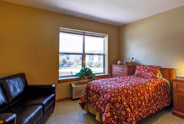 A cozy bedroom with a single bed covered in a red patterned bedspread, a wooden nightstand with a lamp, a wooden dresser, a black leather loveseat, and a large window showing an outdoor garden area.