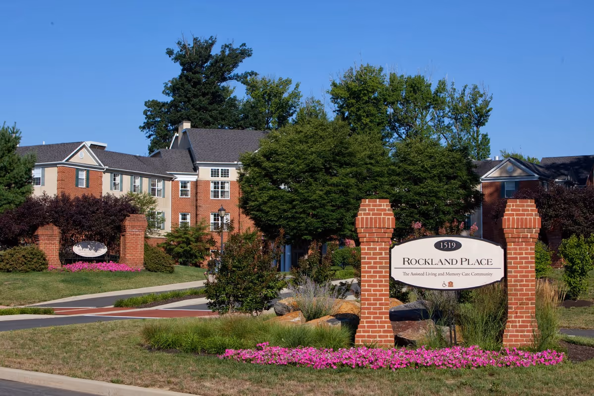 Brick entrance sign reading 'Rockland Place' in front of a multi-story red-brick assisted living building surrounded by trees and flowers.