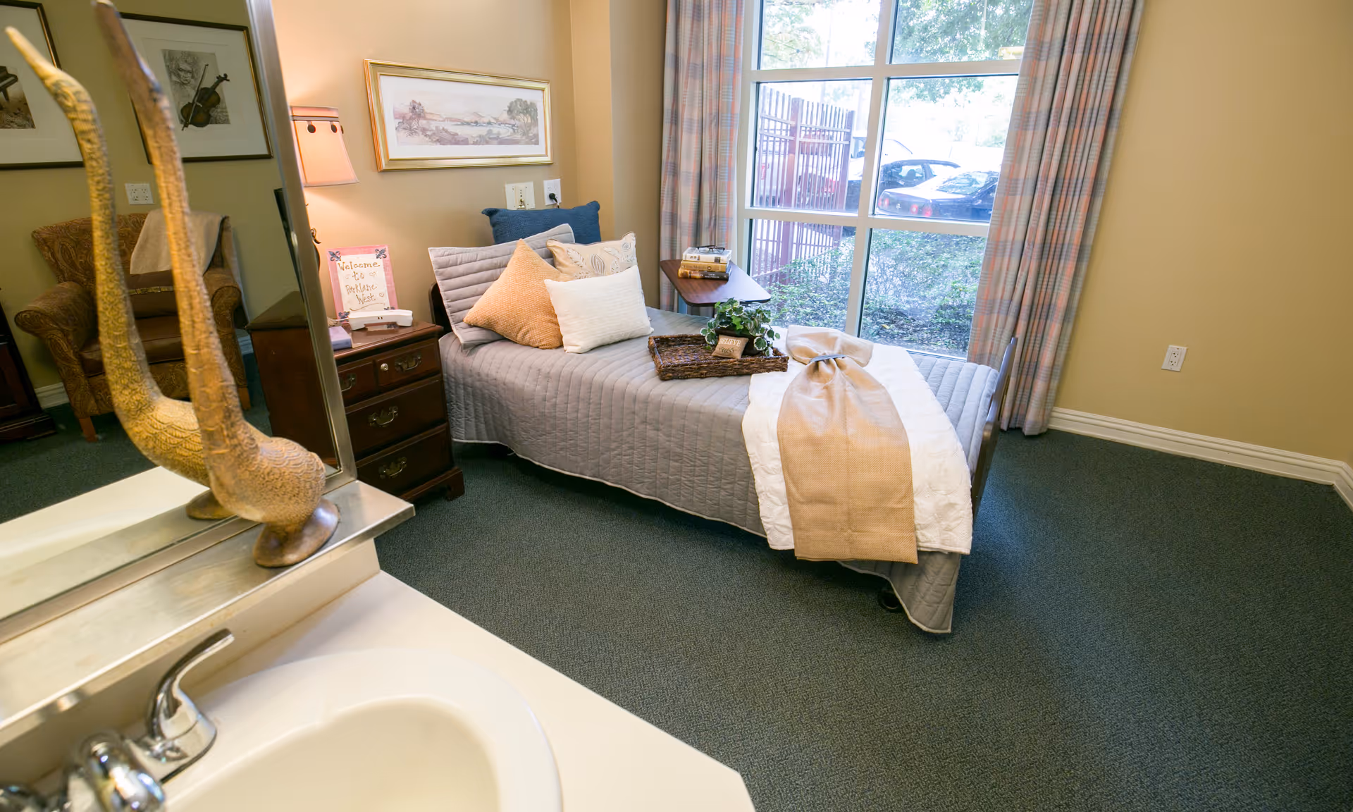 A cozy bedroom in a senior living facility with a single bed covered in a gray quilt and multiple pillows. A tray with a plant and a small sign is placed on the bed. There is a large window with plaid curtains letting in natural light. A wooden nightstand with a lamp and a welcome sign is next to the bed. A comfortable armchair is visible in the background, and a sink with a faucet is in the foreground.