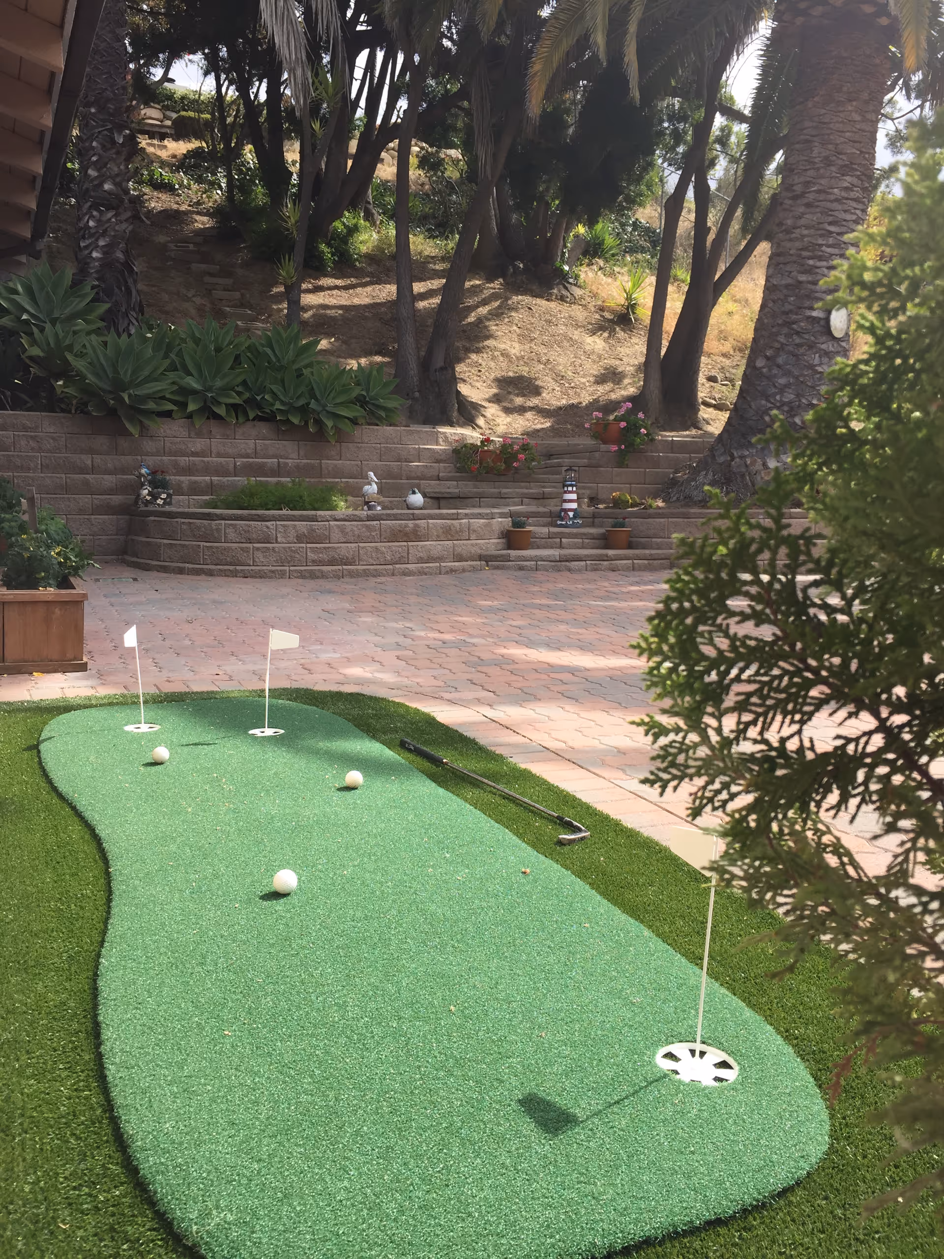 Small outdoor putting green with flags, golf balls and a putter in a paved courtyard with terraced planters and trees.